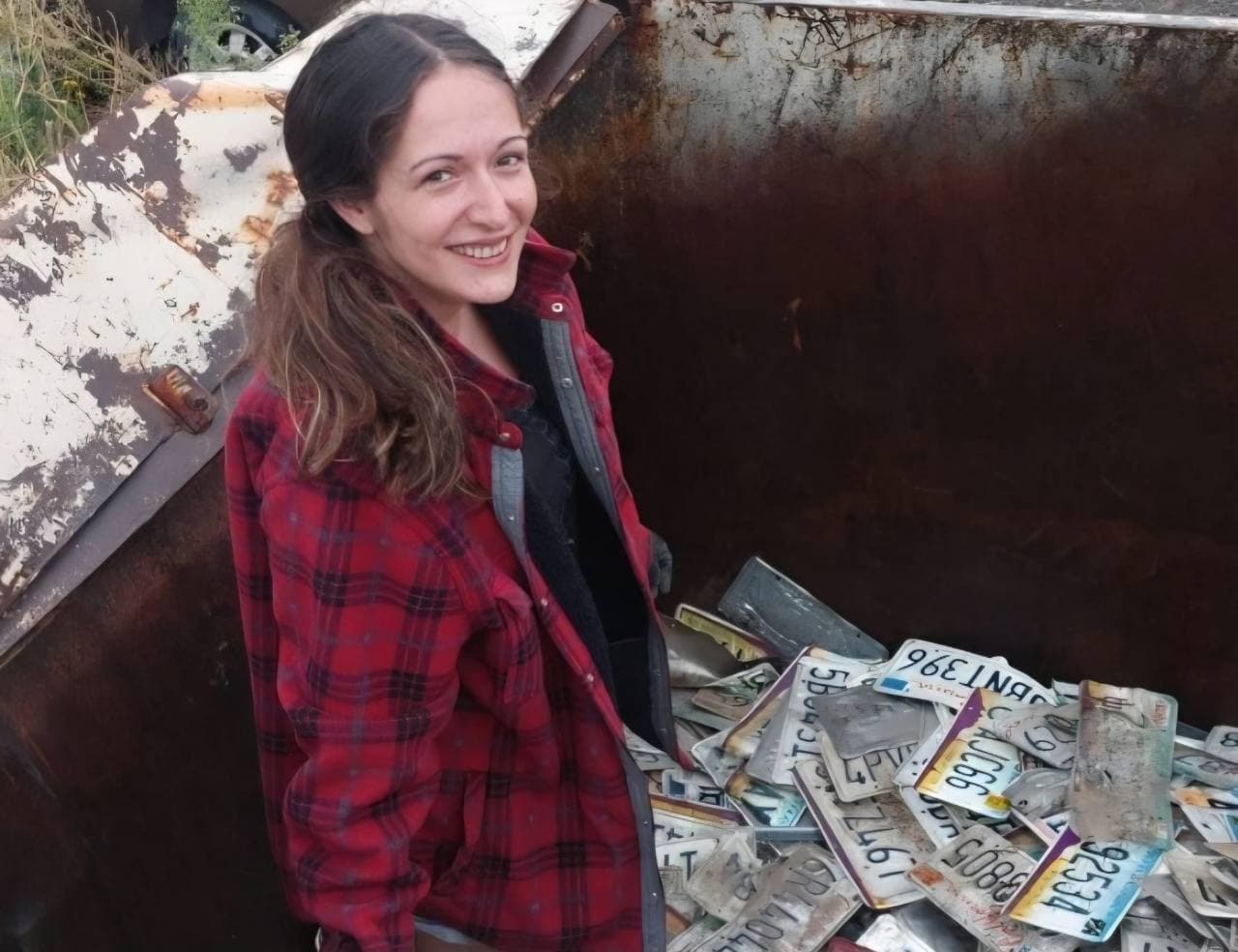 Lächelnde Sammlerin in roter Jacke neben alten Nummernschildern in einem Metallcontainer.